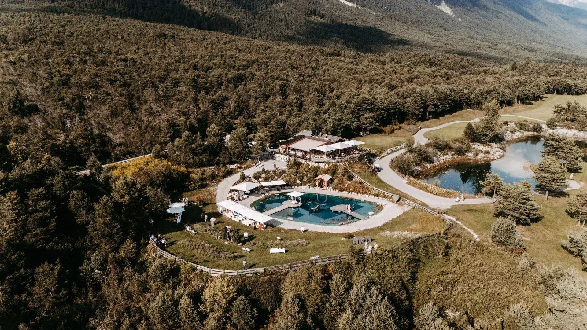 Luftaufnahme der Stöttlalm mit türkisem Badeteich und Almhütte eingebettet in den Wald am Mieminger Plateau