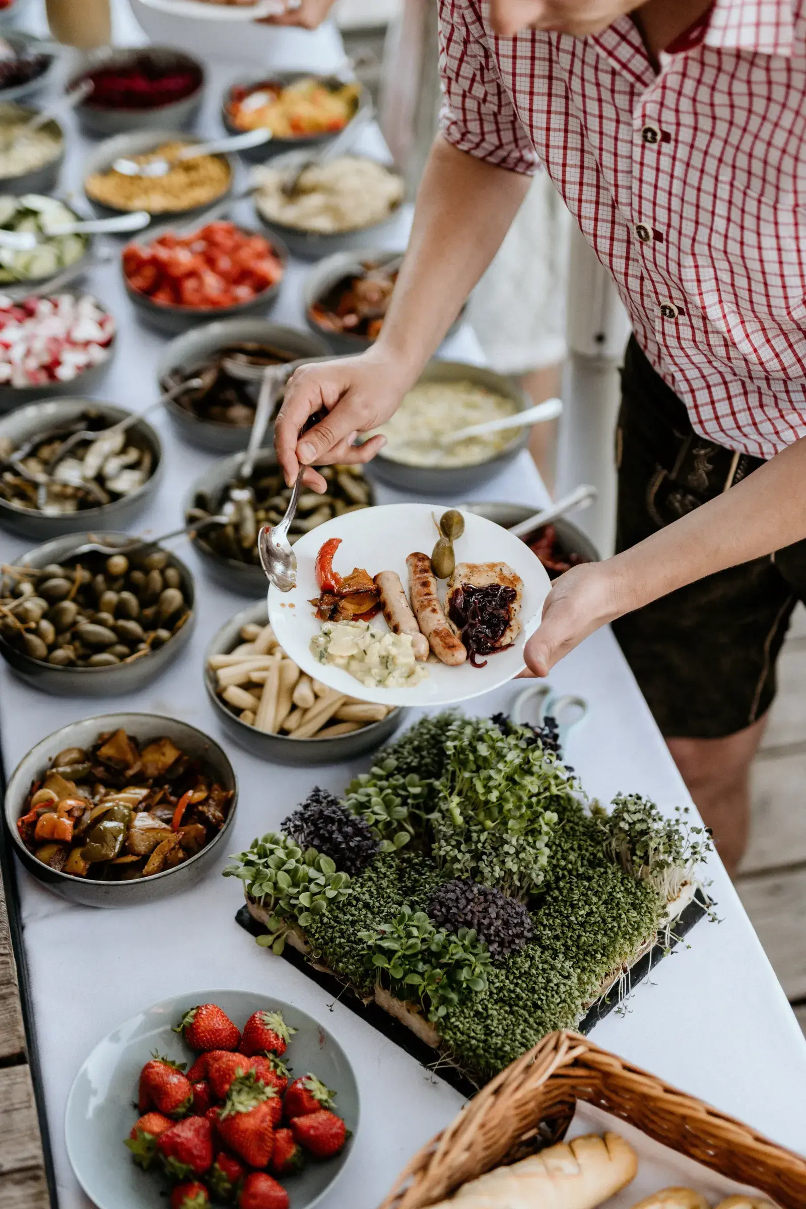 Gast in Lederhose richtet sich am Labestationen-Buffet der Stöttlalm einen Teller mit Bratwurst und Antipasti an