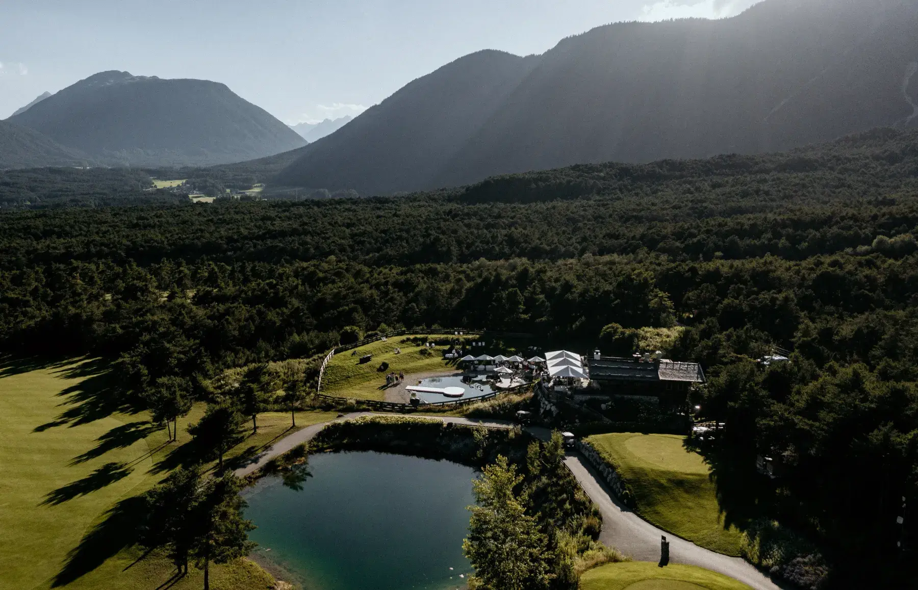 Luftaufnahme der Stöttlalm zwischen Golfplatz, Bergsee und bewaldeten Hängen vor dem Bergpanorama des Mieminger Plateaus